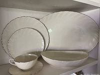 Ivory Lenox Weatherly porcelain pieces stacked showing large serving platter, medium serving bowl, two dinner plates, teacup with saucer in front, all detailed with silver trim and leaf embossing.