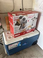 Two boxed Nesco stainless steel coffee urns stacked on a Coleman wheeled cooler in a garage setting.