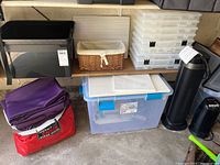View of black plastic storage bin, multiple clear plastic organizers stacked, wicker basket, folded tablecloths or fabric items, large clear plastic bin, and black tower heater and desk fan on the floor.