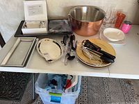 Table top display of various party trays, dishes, drinkware, and utensils including metal trays and a copper-toned punch bowl