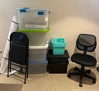 Photo showing stacked plastic storage totes with lids, small turquoise basket, black storage boxes, black folding chair, and black mesh rolling office chair on carpeted floor.