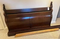 Wooden headboard and footboard with curved sleigh design in dark wood stain laid on carpeted floor.
