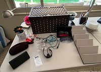 Overview of lot with large woven basket, Fitbit activity tracker with charging cable, assorted pens and pencils, sunglasses in cases, and sunglass wall display stand