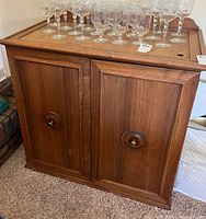 Front view of vintage wooden cabinet showing two paneled doors with round knobs and glassware on top (not included).
