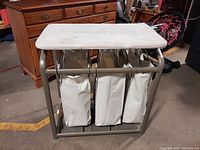 Front view of 3-section laundry hamper with ironing board surface on top showing cloth bags and metal frame.