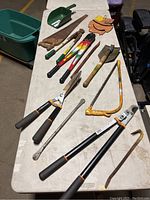 Wide view of assorted tools on a white table showing bolt cutters, pruners, saws, a scoop, and a leather pouch.