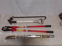 Top view of all four tools displayed parallel: metal drywall hook, large red-handled bolt cutter, heavy-duty door breacher tool with telescopic handle, and metal crowbar