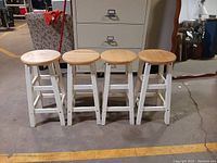 Front view of four wooden barstools with natural round seats and white painted legs placed side by side on a concrete floor.