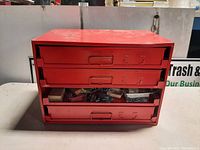 Front view of red metal cabinet showing three drawers with contents inside, including small boxes and hardware.
