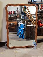 Front view of large mirror with wood frame exhibiting wavy contour, showing scratches on mirror surface and polished wood finish.