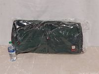 Wilson small rolling carry-on bag, wrapped in factory plastic packaging, displayed standing upright next to a water bottle for scale.