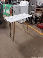 Small sofa table with white marble-patterned wood top and four gold hairpin steel legs shown from front angle in warehouse setting.