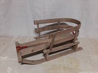 Front and side view of vintage children's wooden sled showing curved backrest, wooden slats, and metal runners with red brackets.