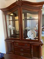 Front view of Ashley cherry wood China cabinet showing curved decorative top with carved detail, glass display doors with glass shelves, and drawers with side cabinets below