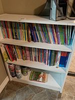 Books on a white bookshelf showing colorful spines of children's books stacked in two rows on three shelves, with various titles visible.