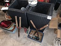 Two large dark woven deep square garden pots with gardening hand tools including shears and hoe in front, small pots stacked on left side.