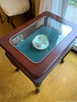 Top view of the side table showing glass inset and wooden frame with a cup and saucer inside the cabinet for scale.