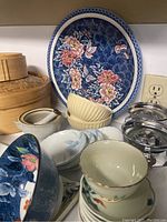 Porcelain bowls and plates with detailed blue and red floral and butterfly patterns, cream ceramic bowls, and stainless steel steam baskets shown stacked on a shelf