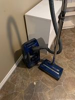 Blue Panasonic canister vacuum cleaner standing on a tiled floor next to a white cabinet. Shows the unit with hose, chrome wand, and broom brush head attachment.