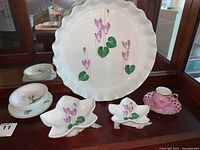 Photo showing large scalloped-edge porcelain tray with purple Cyclamen flowers, two flower-shaped dishes, small white plates with green floral details, and a pink iridescent teacup with gold trim.