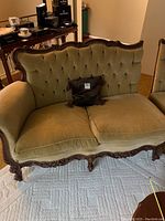 Front angled view of olive green velvet loveseat with carved wooden frame, showing slight staining and seat cushions.