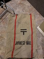 Photo of a vintage Japanese mail bag laid flat on tiled floor showing front side with 'JAPANESE MAIL', red stripes, 'T' symbol and number 72 markings.