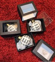 Three white Christmas ball ornaments covered in repeated 'Blah Blah Blah' text in black script, each in an open black square box with clear plastic lid, resting on red carpet.