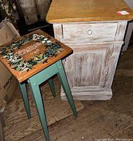 Photo showing solid wood side cabinet with drawer and door, distressed white with natural wood top, and custom painted plant stand with floral decorative top and green legs beside it.