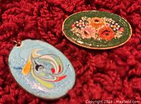 Photo showing both the enameled pendant (blue with abstract enamel art) and the micromosaic pin (oval with floral design) on red fabric