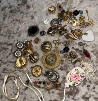 Assorted silver-tone and gold-tone jewelry laid out on grey surface including earrings, pins, pendants and necklace with porcelain floral pendant.