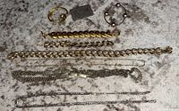 Several pieces of mixed metal tone jewelry including a long silver chain necklace, gold-tone bracelets, black beaded bracelet, and a ring, all laid out on a gray surface.