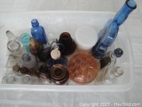 Top view of assorted antique bottles in clear plastic bin including cobalt blue violin flask, amber bottle, and butter pat press.