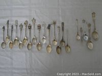 Full view of 20 assorted silver metal collector spoons arranged in a row showing the variety of designs on handles and bowls.