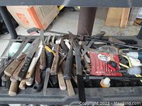 Wide shot of assorted hand tools including several kitchen knives, files, screwdrivers, and nut drivers arranged on a workbench.
