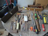 Wide view of assorted hand tools including screwdrivers, pliers, scissors, an antique square, and small hand tools laid out on a table.
