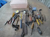 Top-down photo of an assortment of hand tools on a round wooden table shown with a plastic storage box, various pliers, screwdrivers, clamps, combination square, scissors, tape measure and other tools.
