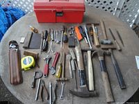 Photo showing miscellaneous hand tools laid out on a round table: multiple hammers (finish, claw, mallet, tacking), various screwdrivers, nut drivers, adjustable wrench, tape measure, files, pliers, leather pouch, and miscellaneous small hand tools.