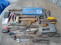 Full overview of assortment of hand tools laid out on a round wooden table including staple gun, screwdrivers, pliers, wrenches, hammers, level and others.