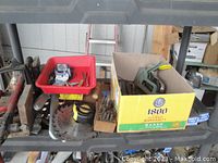 Shelf with assorted hand tools including a red tray containing drill bits and hangers, a box holding staple guns, a hand saw, clamps, tape measure, files, and a circular saw blade.