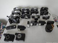13 film camera bodies lined up behind an assortment of flash units and a power winder on a table