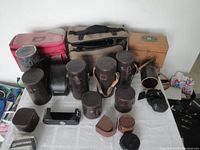 Wide view showing an assortment of vintage leather lens cases, camera cases, and two fabric camera bags laid out on a white surface.