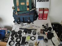 Two camera bags on top of table: one green Canon fabric bag with leather and beige strap details, one dark Tamrac bag below. Various camera lenses, lens caps, lens hoods, small accessories laid out flat around the bags. Two red NY Institute of Photography volumes visible behind the bags. Vivitar camera flash included in items.