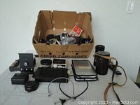 Overview photo of assorted camera accessories including adapters, filters, leather cases, and mounting components arranged on table with a cardboard box in the background
