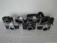 Front view of eight assorted vintage 35mm film camera bodies from Nikon, Canon, Olympus, and Minolta arranged on a table, showing front lens mounts and tops of cameras.