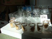 Front view of seven United States Navy shot glasses with gold rims and insignia, along with seven cut crystal stemware glasses on white surface.