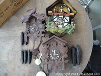 Overview of all three cuckoo clocks with weights and chains laid on a wooden surface.