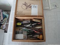 Open wooden box with assortment of hand tools visible including pliers, scissors, tweezers, and small screwdriver set