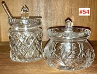 Two crystal lidded jam pots placed side by side on a wooden surface, showing detailed cut-glass and etched patterns. One pot includes a spoon.
