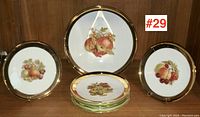 Full view of 7 piece dessert service with fruit motif and gold rims, arranged with serving plate in center and six smaller side plates around it.