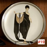 Front view of an Art Deco style porcelain decorative plate featuring a woman in a flowing black dress with gold and blue detailing, standing against a white background with a gold geometric border.